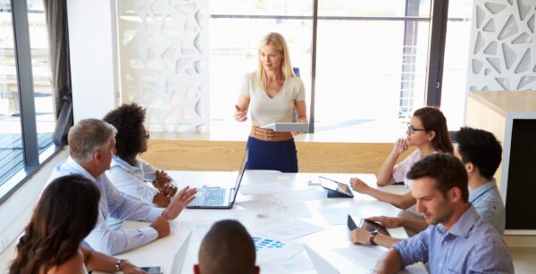 A professional woman in business attire speaking to a group of colleagues seated around a conference table, engaged in discussion.